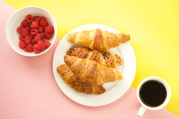 Coffee cup, fresh croissants and raspberries on yellow and pink background