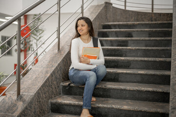 Portrait of charming indian girl reading a book standing on stairs indoors Pretty young student with book on her hands preparing for exams in the library or