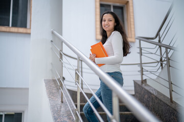 Portrait of charming indian girl reading a book standing on stairs indoors Pretty young student with book on her hands preparing for exams in the library or boo