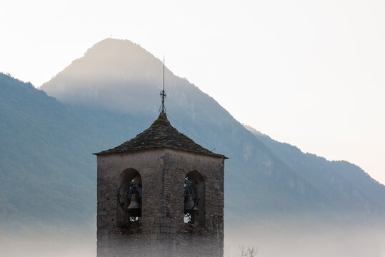 Romanesque bell tower emerges from the fog with mountains in the background and the sun about to rise. Bell tower (12th century ) of the Abbey of San Gemolo in Valganna, province of Varese, Italy 