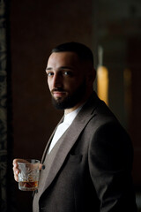 a man with short hair and a beard, wearing a brown three-piece suit. He is standing by the window, holding a glass of drink in his hand.