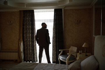 room with dim lighting. In the center of the frame, a man in a suit looks out the window. He stands...