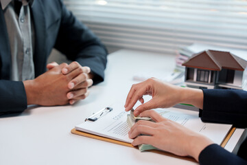 Real estate agent counting us dollar banknotes near contract agreement and house model at office desk, discussing a contract to sell or buy house