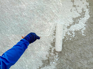 The painter using to hands holding is using a paint roller to apply white paint on the concrete wall.