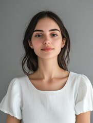 Fototapeta premium Beautiful young woman with long dark hair wearing a white blouse, standing against a soft grey background, showcasing natural beauty and serene expression.