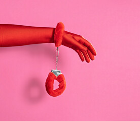 Female hand in red gloves with red fluffy handcuffs on pink background.