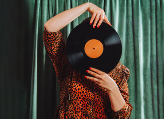 Woman in vintage leopard print dress cover her face with vinyl record on green curtain background