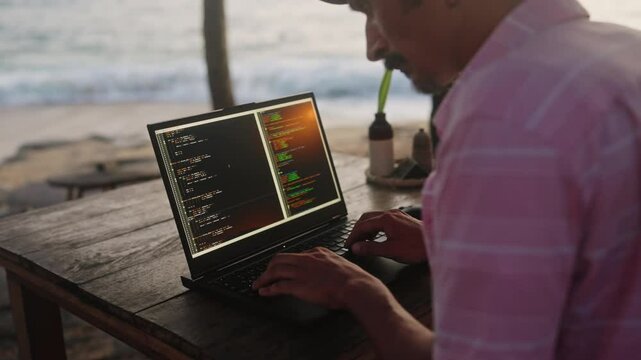 Web developer programs at oceanfront beach cafe at sunset. Male typing, edits code on laptop screen, enjoys tropical seaside view. Remote work combines with relaxing coastal ambiance. Slow motion.