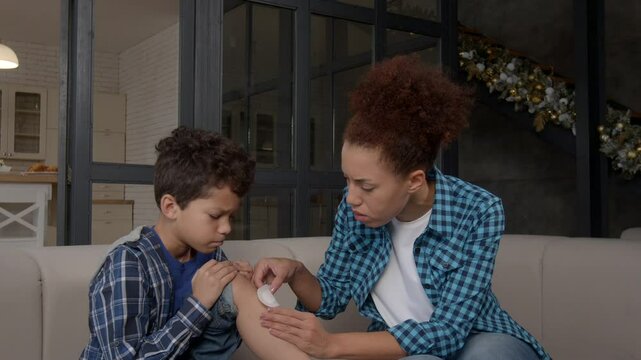 Portrait of caring attractive African American mother providing medical first aid, treating injured knee of school age black son and applying adhesive bandage on wound at home.