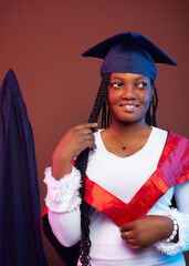 A young woman in a white gown and red cape holds her hair with one hand, smiling proudly. The graduation gown is behind her, emphasizing her achievement.