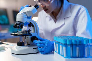 Scientist adjusting microscope while wearing gloves, lab coat, and safety glasses with test tubes...
