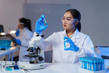 Asian female scientist wearing lab coat and gloves comparing two test tubes with blue liquid while...
