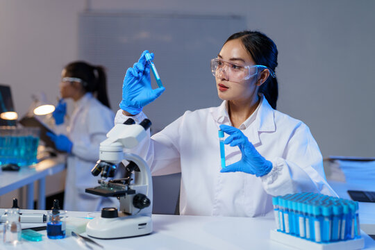 Asian female scientist wearing lab coat and gloves comparing two test tubes with blue liquid while sitting at table with microscope in modern laboratory and colleague working in background