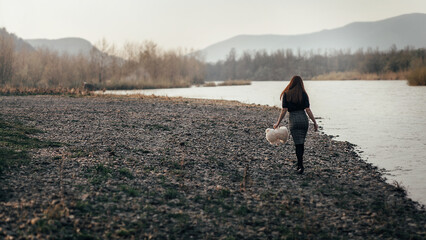 girl walking along the river bank. She is wearing a black sweater and a plaid skirt. The girl is holding in her hand a white plush toy, a bear.