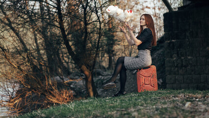 girl with long red hair sitting on a red canister against a background of nature. She's wearing a black blouse and a plaid skirt,