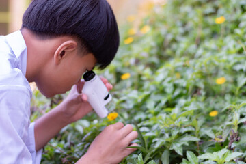 A young teenager holds a magnifying glass to look at plants, leaf, and flowers in school's...