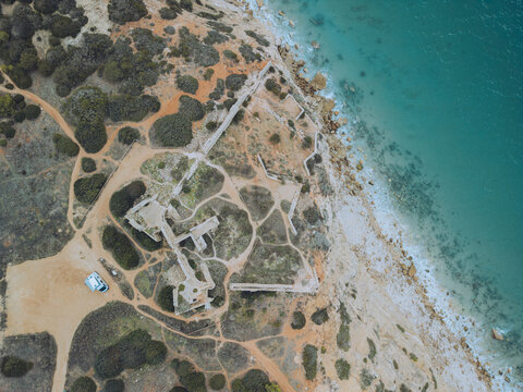 Aerial view of the historic ruins of Forte de Almadena on a rugged cliff by the turquoise ocean, Budens, Portugal.