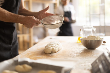 Close up of baker hands dusting raw dough with flour through sieve on kitchen counter in bakery