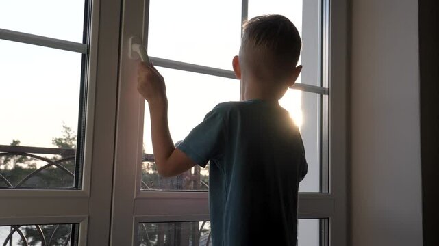 Little boy looks out window at golden hour, watching street in warm evening light. Curious child wants to walk in fresh air. Kids knowledge of world. Sick child is self isolating at home