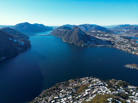 Aerial view of serene Lake Lugano surrounded by picturesque mountains and urban landscape at sunset, Bre sopra Lugano, Switzerland.