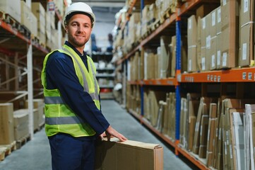 Warehouse worker in blue uniform holding a large cardboard box. Stacks of boxes are visible in the...