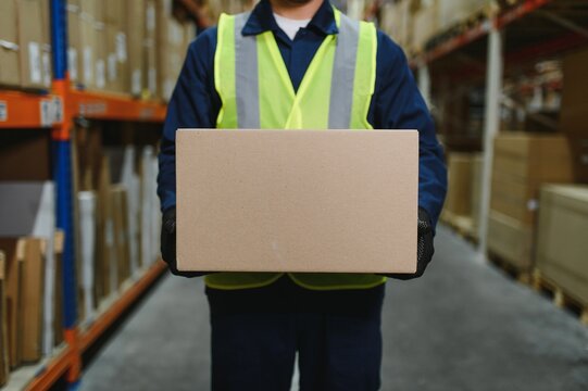 Warehouse worker in blue uniform holding a large cardboard box. Stacks of boxes are visible in the background