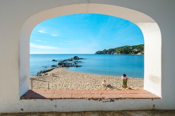 Fototapeta premium People on the beach seen through one of the arches of the promenade of the Picturesque Historical and Tourist City of Calella de Palafrugell, Costa Brava, Catalonia.