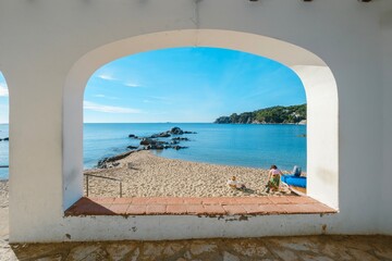 Obraz premium People on the beach seen through one of the arches of the promenade of the Picturesque Historical and Tourist City of Calella de Palafrugell, Costa Brava, Catalonia.