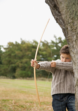 Young boy standing by a tree aiming with a bow and arrow
