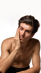 Obraz premium Young attractive man posing, sitting gently holding hand on cheek with cream on forehead against white background. Concept of male beauty and facial care, cosmetology, dermatology. Ad