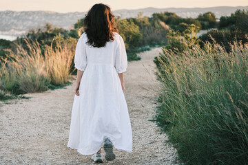 Serene woman in a flowing white dress walks along a tranquil path. Golden hour light bathes the...