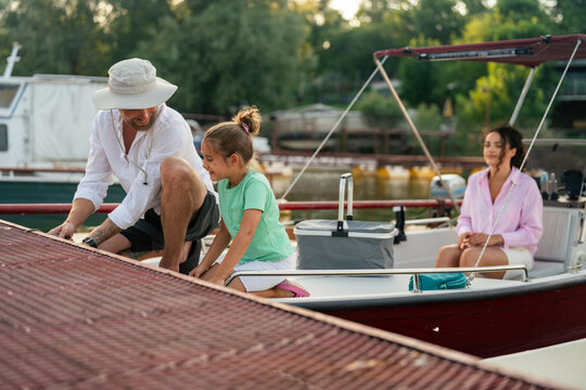 Father and daughter securing a boat to a dock with ropes. They are both kneeling on the boat, and the man is wearing a sun hat and a white shirt. The background shows other boats and a marina.