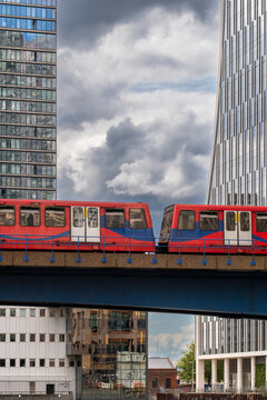A vibrant red and blue train glides across a bridge in an urban setting, surrounded by towering glass buildings under a dramatic sky filled with swirling clouds 