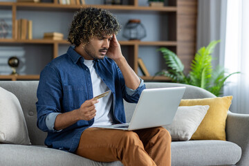 Disappointed and deceived man sitting on sofa in living room with laptop and bank credit card in...