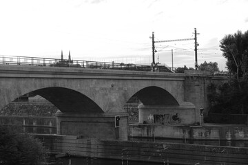 Stone bridge over a river for trains and trams in urban city location with sky and Prague city in the background in black and white 