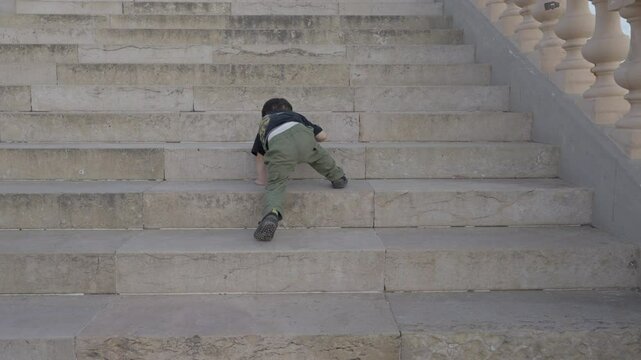 Boy climbs old gray stone stairs, slips and falls gently by himself