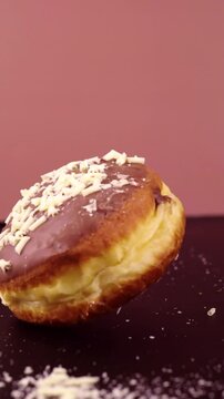 A Berliner donut with a glossy chocolate glaze and white chocolate shavings is dropped in slow motion. The fluffy dough bounces slightly as it lands, scattering chocolate bits on a dark surface.
