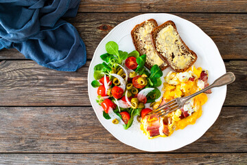Continental breakfast - scrambled eggs with bacon, slices of bread and fresh salad on wooden table