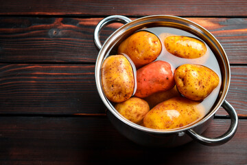Potatoes with peel in a stainless steel pot with boiling water. Cooking dishes from potatoes. On a wooden background. Free space for text.