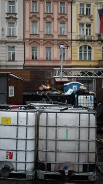 Vertical frame showing large plastic containers and scattered decorations in a city square, with historic facades in the background, marking the beginning of holiday preparations.
