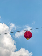 A red Chinese lantern hangs from a wire against a bright blue sky with scattered white clouds.