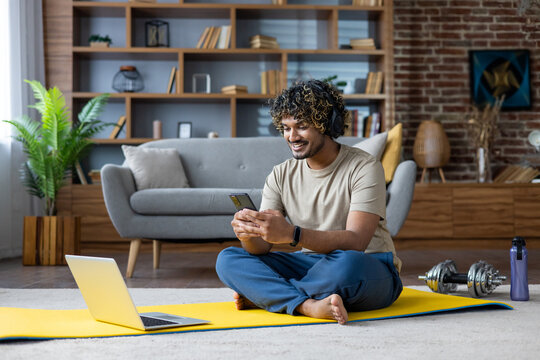 Young athletic man sitting on the floor of the apartment in the living room, on a fitness mat and using an application on his phone, smiling happily. - Powered by Adobe
