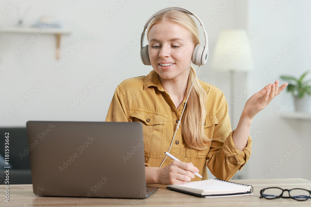 Poster interpreter in headphones taking notes while having video chat via laptop at table indoors