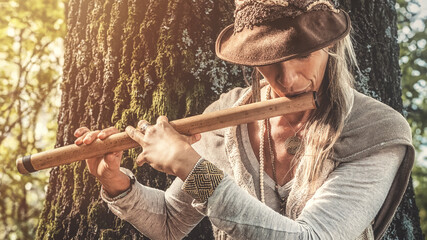 A woman playing a wooden pipe near a tree in the forest