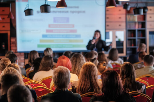 Conference attendees listen to a speaker in a modern venue with presentation slides visible.