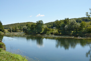 A peaceful scene of the Dobra River near Jaskovo, showcasing its clear waters reflecting the sky and surrounding greenery, a small dam, and an old bridge in the distance
