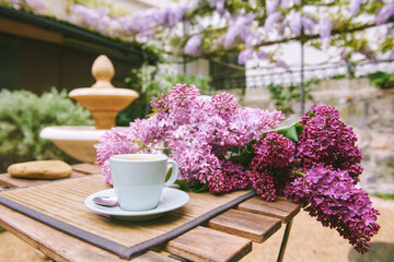 Romantic image of spring lilac bouquet with cup of coffee on the table in outdoor cafe