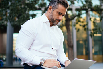 Mature male trader making online business messaging sitting outdoors at urban setting, middle aged entrepreneur using 4g wireless internet connection for uploading web files on modern laptop computer