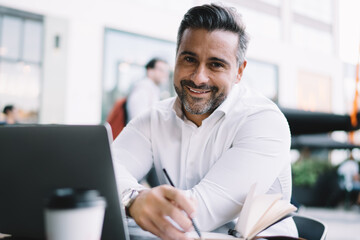 Portrait of cheerful entrepreneur dressed in formal white shirt smiling at camera during time for business planning, middle aged male employer working remotely in sidewalk cafe enjoying lifestyle