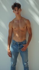 Fototapeta premium A handsome young man stands against a minimalist white background, showcasing his toned physique in fitted blue jeans. The interplay of light and shadow accentuates his strong features, exuding a